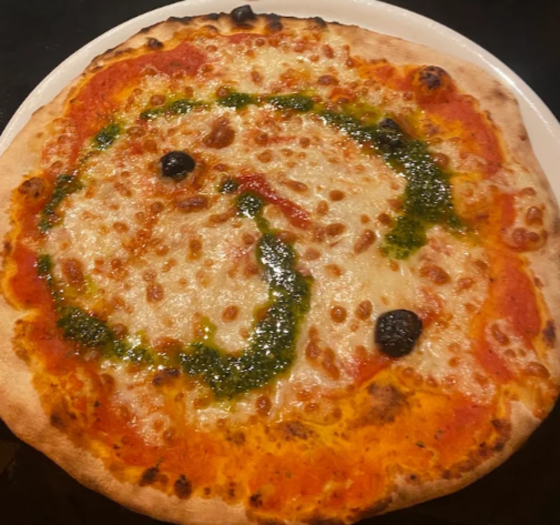 Une pizza sortie du feu de bois, cordon de pesto vert et olives noires sur la mozzarella